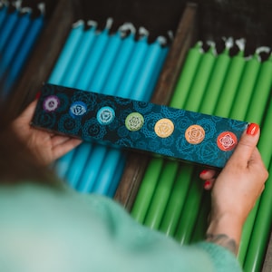 Op de afbeelding: Een doos met zeven chakra-kaarsen met een zwarte achtergrond en kleurrijke chakra-symbolen. De kaarsen zijn in een rij gerangschikt en zijn gelabeld met de namen van de chakra's. De doos wordt vastgehouden door de handen van een persoon.