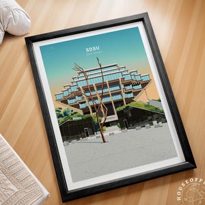 May include: Framed art print featuring a stylized illustration of the San Diego State University (SDSU) library. The print has a black frame and depicts the building with a tree in front. The sky is a gradient of blue and orange.