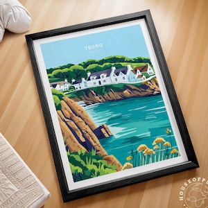 May include: Framed art print featuring a coastal landscape with the text "TRURO CORNWALL". The artwork depicts white buildings, green trees, and a turquoise sea. The print is in a black frame and is displayed on a wooden surface.