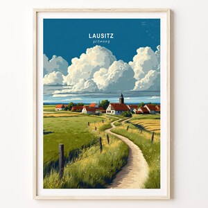 May include: A framed art print of a landscape scene. The artwork depicts a rural village with red-roofed houses, a winding path, and fields of green and yellow. The sky is a deep blue with large, fluffy white clouds. The text "LAUSITZ Germany" is at the top.