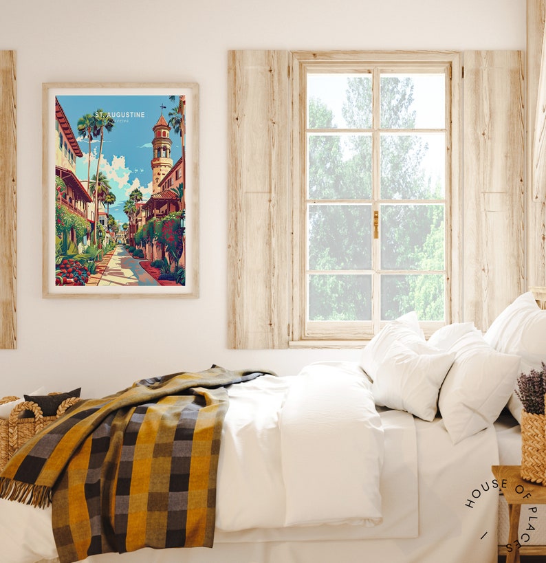 May include: A framed print of a colorful illustration of a street scene in St. Augustine, Florida. The print is hanging on a white wall above a bed with a white duvet and a yellow and black plaid throw blanket.