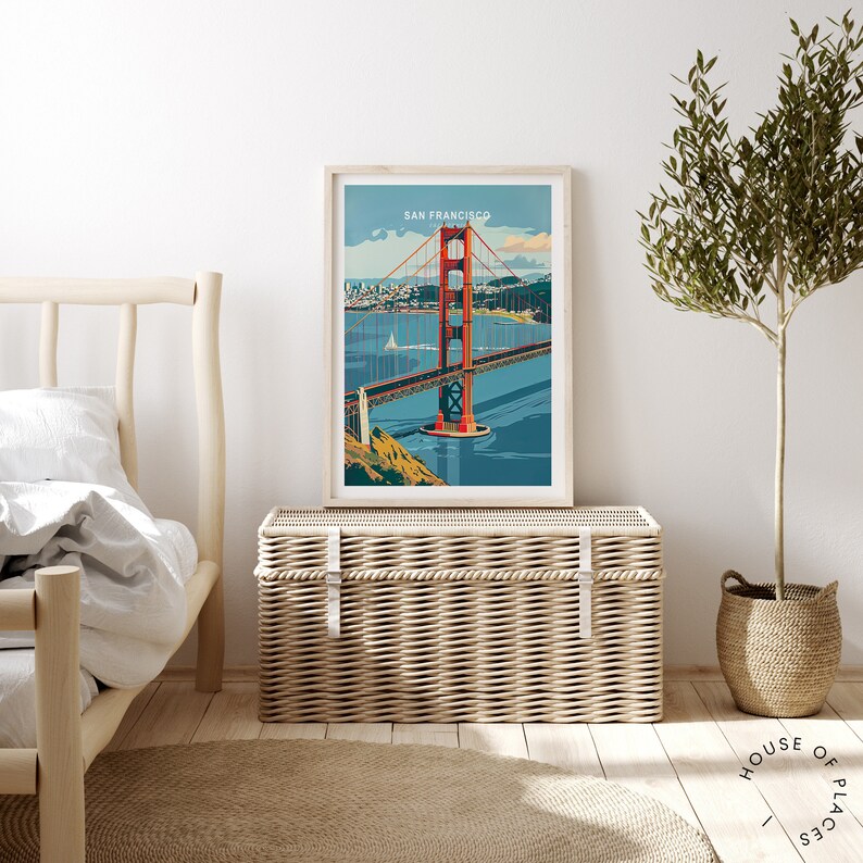 May include: A framed print of the Golden Gate Bridge in San Francisco, California. The bridge is red and orange and is set against a blue sky and water. The print is hanging on a white wall above a wicker storage trunk.