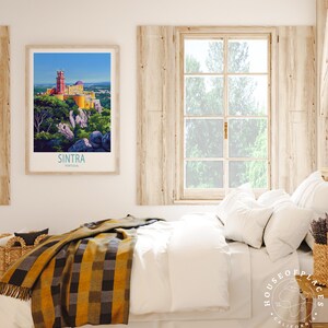 May include: A framed print of Sintra, Portugal, showing a vibrant castle on a hilltop, with the text "Sintra Portugal" below. The print is above a bed with white bedding, a plaid blanket, and a window overlooking a green landscape.