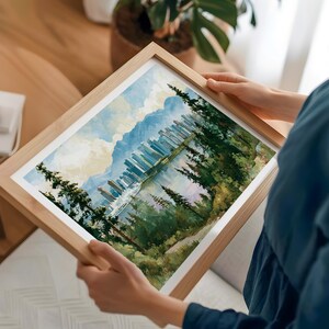 May include: A framed landscape print featuring a watercolour-style painting of a city skyline nestled between dense evergreen forests and distant mountains, with a reflective body of water in the foreground under a soft, cloudy sky.