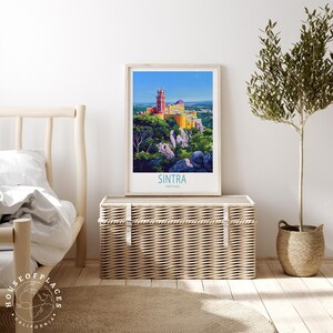 May include: A framed art print depicting Sintra, Portugal, showcasing a vibrant illustration of a castle on a hilltop. The print is in a light wood frame, with the word "Sintra" in teal at the bottom. It's displayed on a woven chest, with a small tree nearby.
