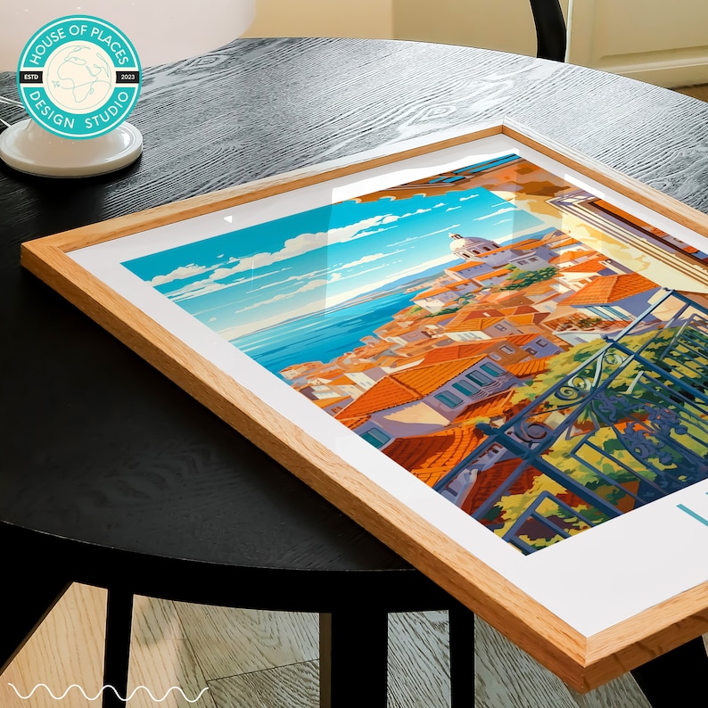 May include: A framed print of a colorful illustration of a cityscape with a blue sky and ocean in the background. The city is built on a hillside and has many red-tiled roofs. The print is in a wooden frame.