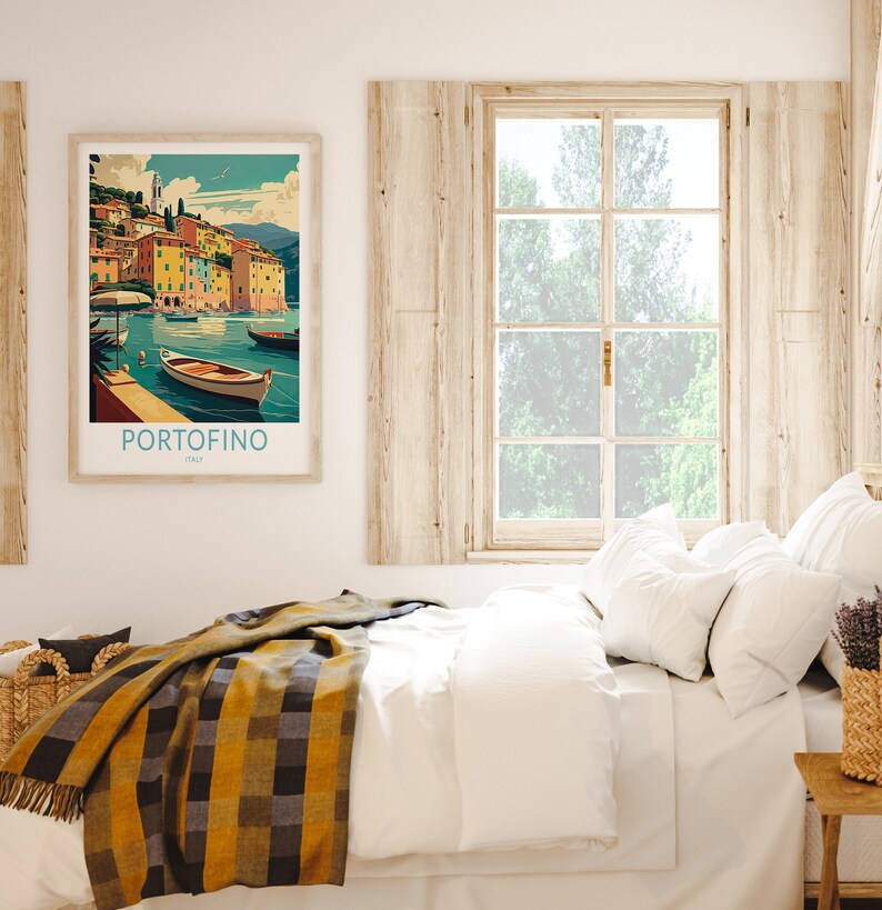 May include: A framed print of a colorful illustration of the Italian town of Portofino, with boats in the water. The print is hanging on a white wall above a bed with white bedding and a yellow and black plaid throw blanket.