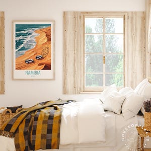 K&ouml;nnte beinhalten: Ein Vintage-Reiseposter mit einem blauen und wei&szlig;en Bild eines Strandes in Namibia, Afrika. Das Poster ist gerahmt und h&auml;ngt an einer wei&szlig;en Wand &uuml;ber einem Bett mit einer wei&szlig;en Bettdecke und einer gelb-schwarzen karierten Wolldecke.