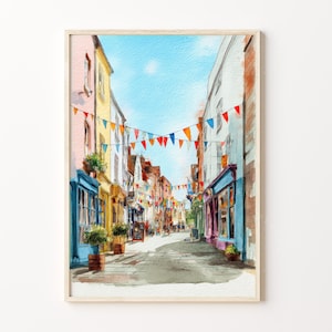 May include: Watercolour painting of a European street scene. Buildings in various colours line the cobbled street, decorated with hanging flags. The sky is a bright blue, and the artwork is framed in light wood.