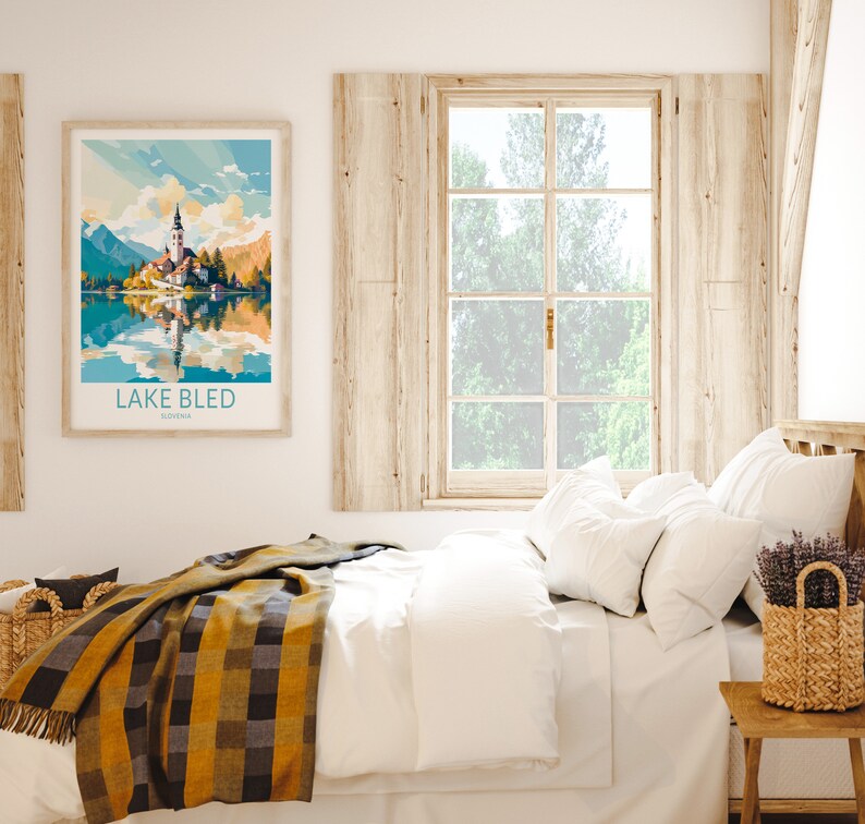 Op de afbeelding: Een wit bed met een geel en zwart geruit deken, witte kussens en een rieten mand met lavendelbloemen op een houten nachtkastje. Een ingelijst afdruk van het meer van Bled, Sloveni&euml;, hangt aan de muur boven het bed.