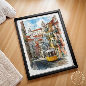 Pu&ograve; includere: Stampa artistica incorniciata di una scena di strada di Lisbona con un tram giallo sui binari, edifici colorati e una chiesa sullo sfondo. L'opera d'arte &egrave; in una cornice nera ed &egrave; appoggiata su una superficie di legno. La stampa &egrave; di House of Places California.