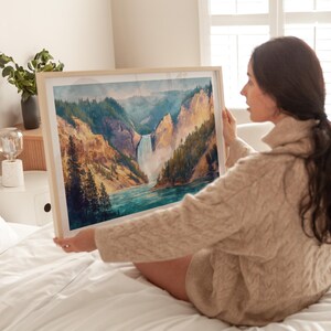 Op de afbeelding: Een ingelijste aquarel schildering van een waterval die in een rivier stort, omringd door kliffen en bomen. Het kunstwerk heeft tinten blauw, groen en bruin. De lijst is licht van kleur.
