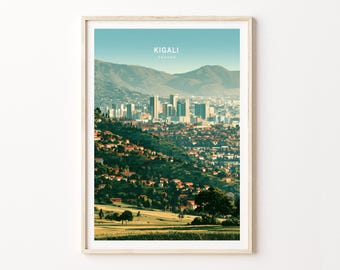 May include: A framed wall art print featuring a panoramic landscape illustration of Kigali, Rwanda. The image displays a city skyline with tall buildings, rolling green hills, residential houses with orange roofs, and mountains under a clear sky.