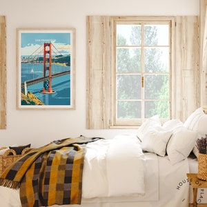 May include: A framed poster featuring a vintage illustration of the Golden Gate Bridge in San Francisco, California. The poster is hanging on a white wall above a bed with a white duvet and a yellow and black checked throw blanket.