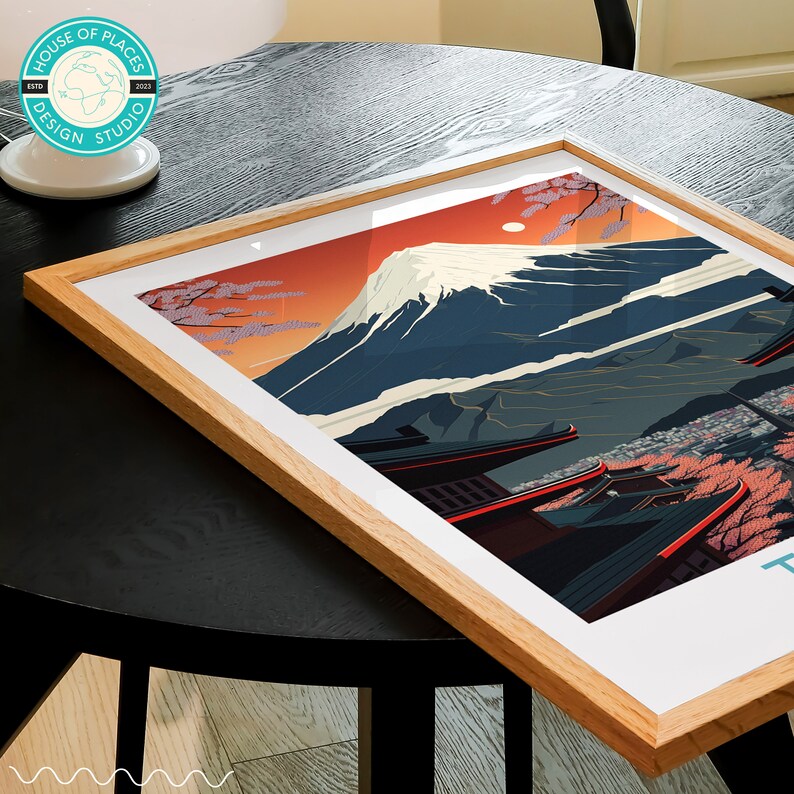Pu&ograve; includere: Una stampa incorniciata del Monte Fuji, una famosa montagna giapponese, con un cielo rosso e fiori di ciliegio in primo piano. La stampa &egrave; in una cornice di legno e ha il testo "House of Places Design Studio" in un cerchio in basso.