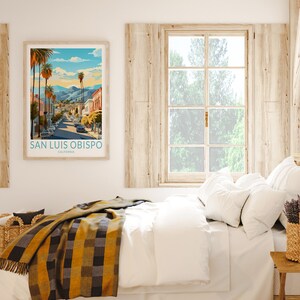 May include: A framed print of a vintage travel poster for San Luis Obispo, California. The poster features a colourful illustration of a street scene with palm trees and buildings. The poster is hanging on a white wall above a bed with white bedding and a yellow and black plaid throw blanket.