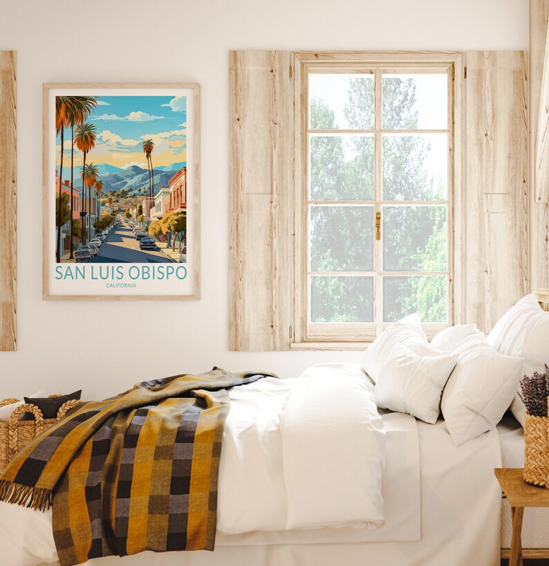 May include: A framed print of a vintage travel poster for San Luis Obispo, California. The poster features a colourful illustration of a street scene with palm trees and buildings. The poster is hanging on a white wall above a bed with white bedding and a yellow and black plaid throw blanket.