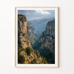 May include: A framed landscape photograph of a deep canyon with towering rock formations. The canyon walls are a mix of tan and gray, with green vegetation. The sky is a pale blue with some clouds, and the overall scene is bathed in sunlight.