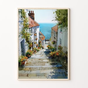 May include: Watercolour painting of a charming European village street leading to the sea. White buildings with red-tiled roofs and flower boxes line the cobbled path. The artwork is framed in a light wood.