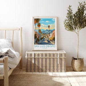May include: A framed poster featuring a colourful illustration of a street scene in San Luis Obispo, California. The poster depicts a blue sky with white clouds, palm trees, and buildings. The text on the poster reads "San Luis Obispo, California".