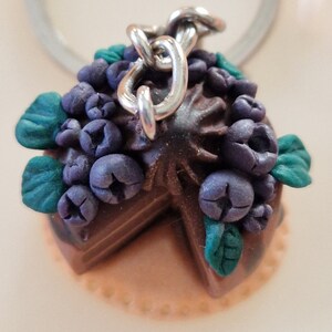 May include: A chocolate cake keychain with a slice missing, decorated with purple blueberries and green leaves. The cake is attached to a silver chain.