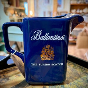 May include: A deep blue ceramic jug featuring the white script text Ballantine's, a gold-coloured heraldic crest, and the white text THE SUPERB SCOTCH. The jug has a square-shaped body, a side handle, and a wide pouring spout at the top.