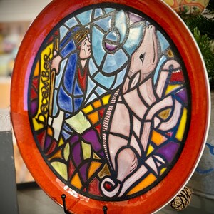 May include: A decorative ceramic plate with a stained-glass design. The plate has a red border and a central image with a figure, a donkey, and the word "December". The artwork uses blue, purple, yellow, and orange colours.