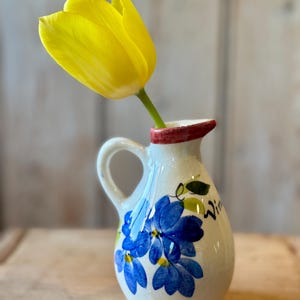 May include: A small, white ceramic jug with a handle, decorated with blue floral designs and the word "Wine". A bright yellow tulip is placed inside the jug. The rim of the jug is red.