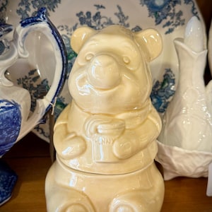 May include: A cream-coloured ceramic jar shaped like a seated bear holding a small pot with the word HUNNY embossed on the front. The bear has rounded ears, a smooth glossy finish, and a two-piece construction with a removable lid at the waist.