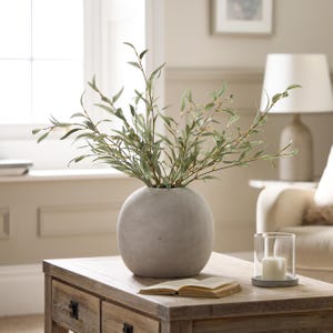 May include: A spherical, grey concrete vase holding green foliage is displayed on a wooden coffee table. A glass candle holder with a lit white candle and an open book are also on the table. A lamp and a window are in the background.