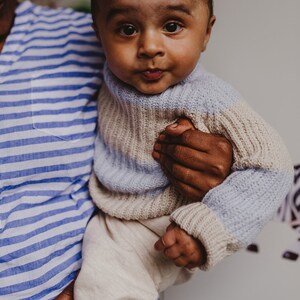 May include: A baby wearing a light blue and beige striped sweater is being held by an adult. The baby is looking directly at the camera with a curious expression.