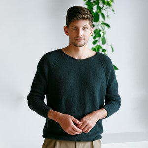 May include: A man wearing a dark green sweater with a round neckline. He is standing in front of a white wall with green plants in the background.