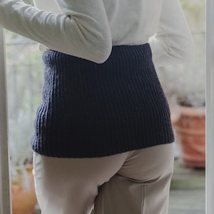 May include: A woman wearing a white long-sleeved shirt with a black knitted belt. She is standing in a doorway and looking out.