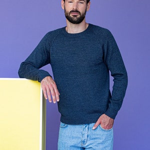Peut inclure: Un homme portant un pull bleu foncé et un jean bleu clair se tient devant un mur violet. Il s'appuie contre un objet rectangulaire jaune et blanc.