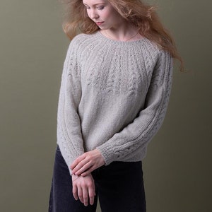 May include: A light grey knitted jumper with a cable knit pattern on the front and sleeves. The jumper has a round neckline and a relaxed fit.
