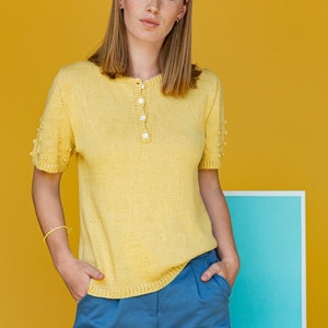 May include: A woman wearing a yellow knitted short-sleeved shirt with a button-up collar and blue pants. She is standing in front of a yellow wall with a blue rectangle behind her.