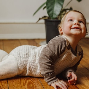 Puede incluir: Un bebé con un mono de punto beige y leotardos blancos está gateando en un suelo de madera. El bebé está mirando hacia arriba y sonriendo.