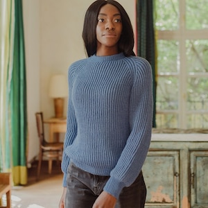 以下が含まれることがあります： 丸首と長袖の青いニットセーターを着た女性。窓と木製のドアのある部屋に立っています。