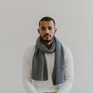 May include: A man wearing a white long-sleeved shirt with a black and white striped scarf around his neck. He has a short beard and is looking directly at the camera.