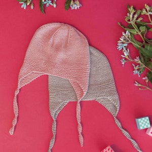 May include: Two knitted baby hats with ties, one pink and one gray, on a red background with wooden blocks.