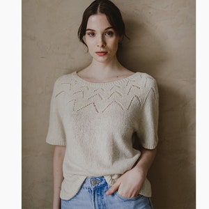 May include: A white knitted sweater with a delicate openwork pattern around the neckline. The sweater has short sleeves and a relaxed fit.