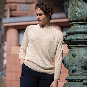 Könnte beinhalten: Ein hellbeige Rundhals-Pullover mit einem strukturierten vertikalen Muster. Der langärmlige Strickpullover wird mit dunkelblauen Hosen kombiniert. Die Person steht neben einem dekorativen grünen Metallpfosten.