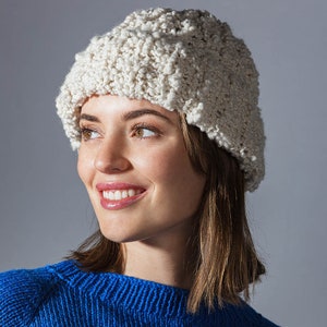Könnte beinhalten: Eine weiße Strickmütze mit texturierter Oberfläche. Die Mütze wird von einer Person mit braunen Haaren und einem blauen Pullover getragen.