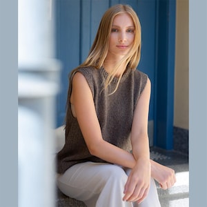 Könnte beinhalten: Eine ärmellose braune Strickweste mit Rippstruktur. Die Weste wird über einem hellen Oberteil getragen und mit einer cremefarbenen Hose kombiniert. Das Model sitzt auf einer Steinstufe vor einer blauen Tür.