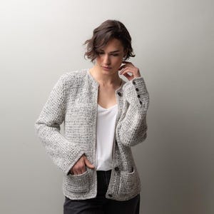 May include: A light gray tweed jacket with a textured weave, featuring a round neckline, two front pockets, and dark buttons. The jacket is worn over a white top and black pants.