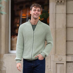 Puede incluir: Un cárdigan verde claro con cremallera y textura de punto acanalado. El cárdigan tiene un cuello alto y una cremallera completa. La persona lleva una camiseta verde oscuro y vaqueros azul oscuro.