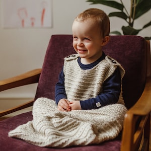 May include: A smiling child wearing a beige knitted sleep sack with button closures on the shoulders. The sleep sack is over a navy blue long-sleeved shirt. The child is seated in a purple armchair with wooden armrests.