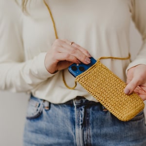 May include: A mustard yellow crocheted phone case with a blue phone inside. The case is attached to a long strap and is being held by a person.