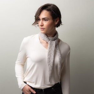 Könnte beinhalten: Ein hellgrauer Schal mit offenem Gewebe und einem zarten, durchbrochenen Muster. Der Schal ist um den Hals drapiert und hängt in zwei langen, spitz zulaufenden Enden herab. Der Schal ist aus einem weichen Material gefertigt.