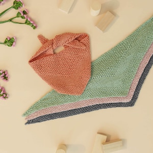 Könnte beinhalten: Vier gestrickte Dreieckslätzchen in verschiedenen Farben: Koralle, Grün, Rosa und Grau. Die Lätzchen sind fächerförmig auf einem hellbeigen Hintergrund angeordnet. Es gibt auch Holzklötze, die um die Lätzchen herum verstreut sind.